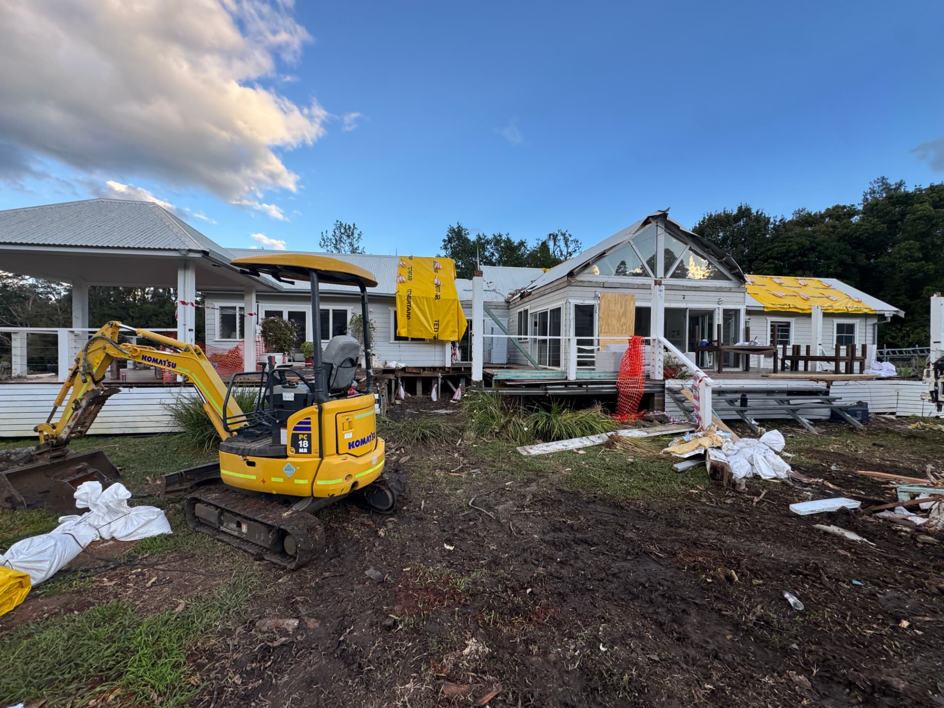 Damaged homestead braced and tarped at dusk with an excavator and safety fencing on site