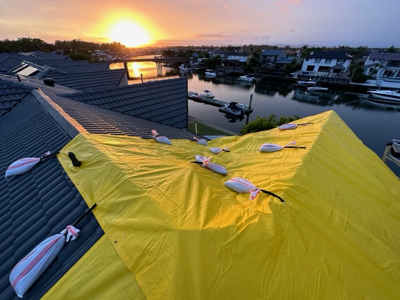 Yellow emergency roof tarp weighted with sandbags over a tiled roof at sunrise