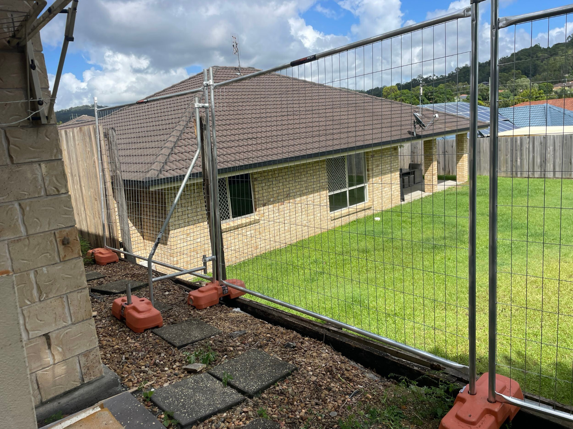 Temporary mesh fence panels on orange feet enclosing the side yard of a tiled-roof brick residence