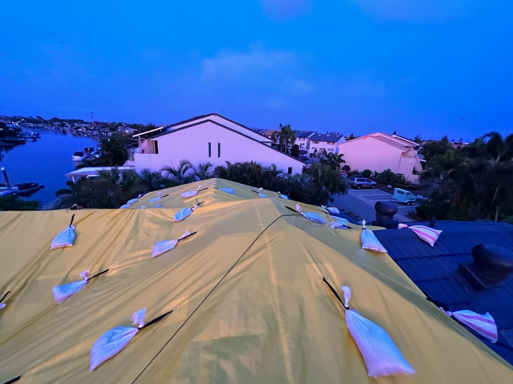 Yellow temporary roof protection ballasted with sandbags across a hip roof at dusk