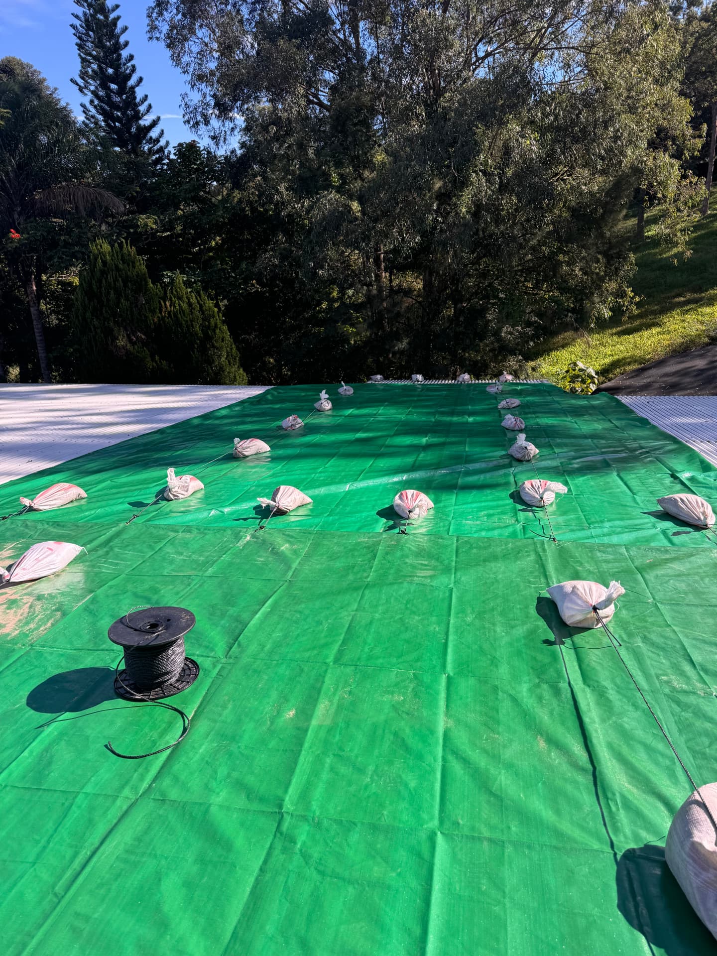 Green temporary roof cover pulled across a metal roof and tied off with sandbags and rope