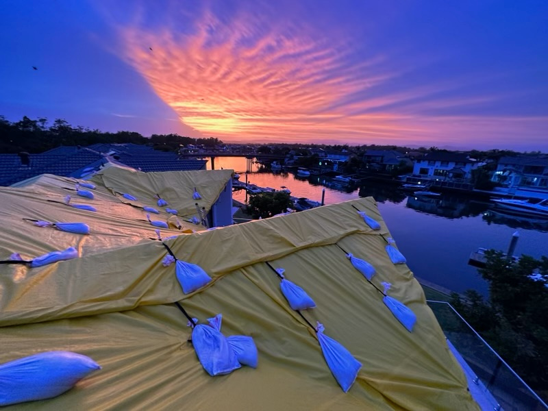 Temporary roof protection tarped and sandbagged across a canal-front roof at sunset