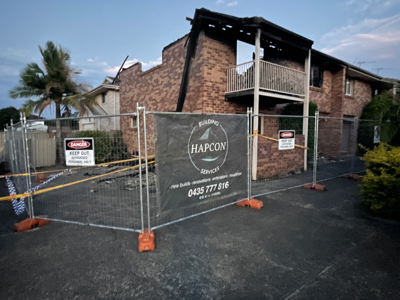 Full temporary fence perimeter with danger signage and hazard tape around a fire-damaged two-storey brick block