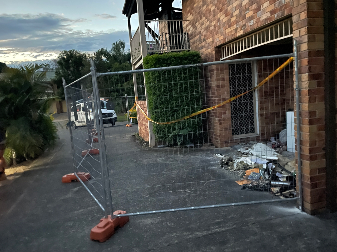 Fence panels and hazard tape closing off the entry of a fire-damaged brick unit at dusk